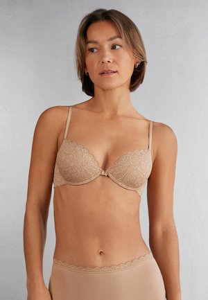 Nude kleur kanten bh met geschulpte randen en verstelbare bandjes. Het ontwerp heeft een strikdetail in het midden en een textuurpatroon.