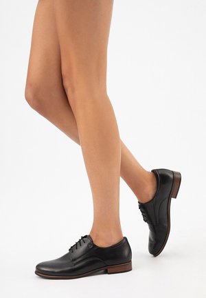 Beine mit schwarzen, aus Leder gefertigten Oxford-Schnürschuhen mit braunen Sohlen, seitlich auf weißem Hintergrund gezeigt.
