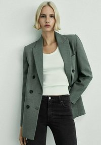 Gray double-breasted blazer with notched lapels and black buttons, worn over a white ribbed top and black jeans, featuring a tailored fit.