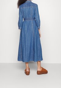 Denim shirt dress with a fitted waist and mid-calf length. Features long sleeves, a collar, and a belted waist. Worn with brown slip-on clogs.