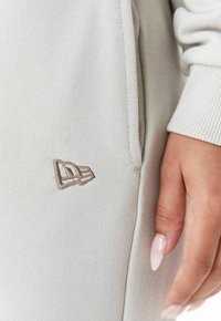Hellegraues Sweatshirtmaterial mit geripptem Textur. Verfügt über ein kleines besticktes Logo in einem Braunschattennah der Tasche. Nahaufnahme.