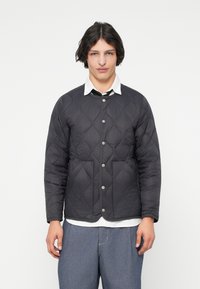 Kviltet svart jakke med trykknapper og rund hals. Den har horisontal søm og to frontlommer. Stillet over en hvit skjorte.