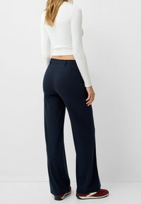 Navy wide-leg trousers with a smooth texture and tailored fit, paired with a fitted white long-sleeve top and red sneakers.