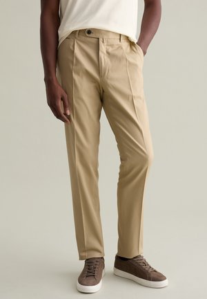 Tan, lightweight trousers with a flat front and single button closure. Worn with brown suede sneakers and a light-colored t-shirt.