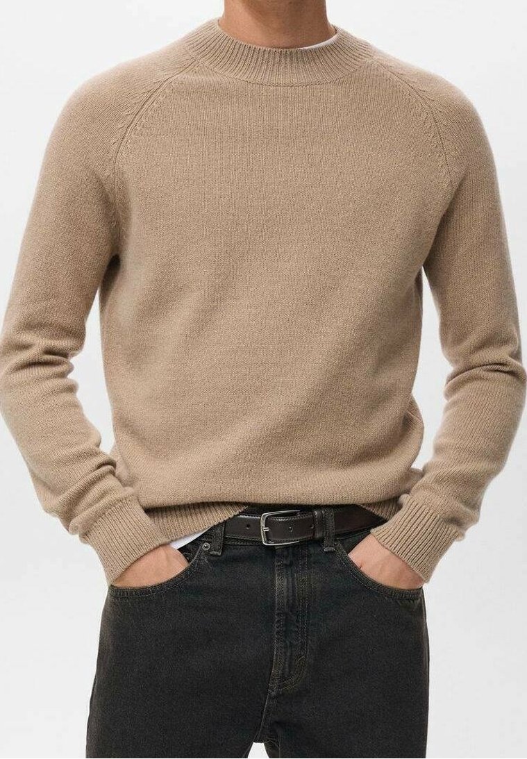 Homme portant un pull en maille beige avec poignets et ourlet côtelés, ceinture noire et jean foncé, mains dans les poches.
