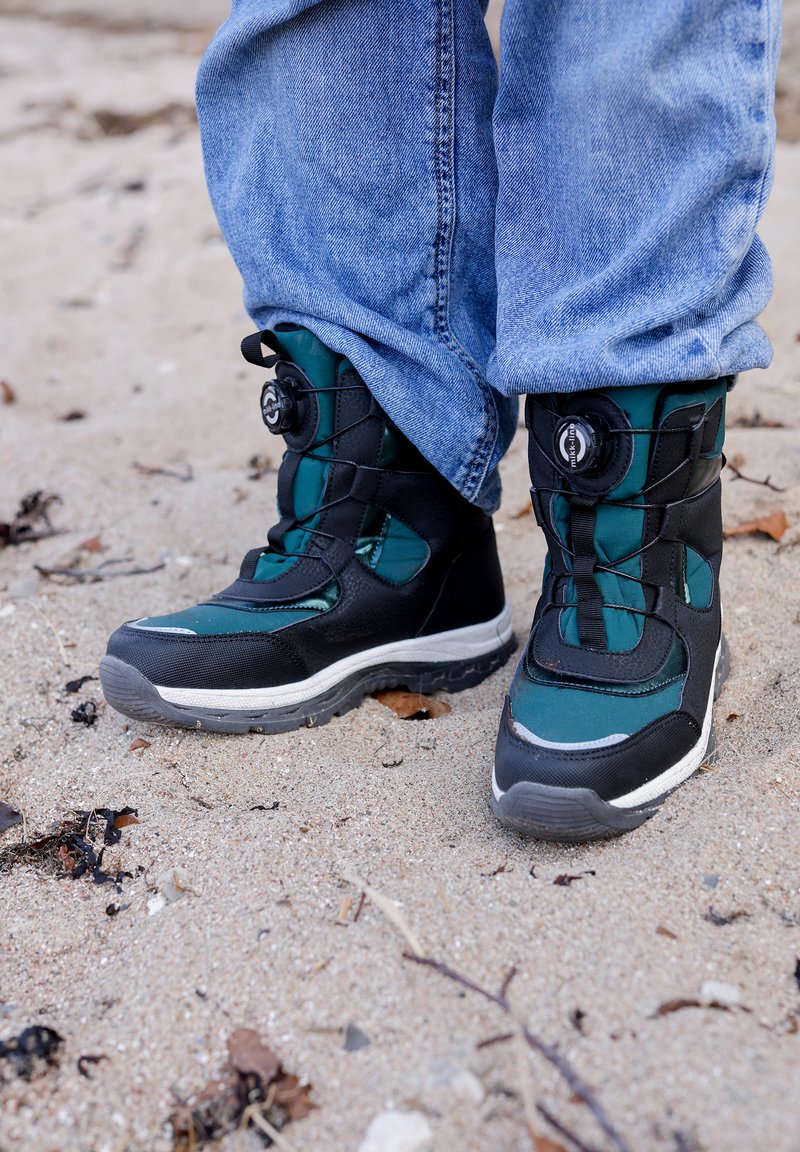 Musta ja petroolinsininen vedenpitävä saappaat, joissa on teksturoitu päälliosa, kaareva kärki ja pyöreä nauhoitusmekanismi, asetettuna hiekkapinnalle.