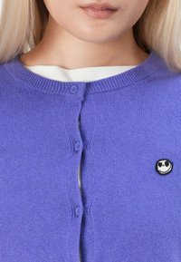Gilet en tricot violet avec un col rond, deux boutons assortis et un petit patch brodé en noir et blanc d'un personnage sur la poitrine.