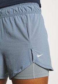 Nike women's shorts in light blue, featuring an elastic waistband, side slits, and a white logo. Made from a smooth, lightweight fabric.