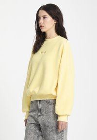 Geel fleece sweatshirt met een relaxed fit, geribbelde manchetten en zoom. Heeft subtiele tonale borduursels op de borst. Draag met grijze jeans.