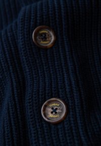 Il tessuto lavorato a maglia blu navy presenta una texture a coste; due bottoni marroni con la parola "OMBRE" stampata in giallo su ciascuno.
