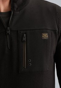 Schwarzes Sweatshirt aus strukturiertem Stoff, mit hohem Kragen und einer Brusttasche mit Reißverschluss. Goldene und schwarze Logoplakette auf der Tasche.