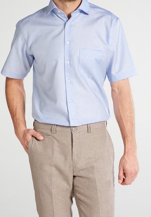 Uomo con camicia celeste a maniche corte con taschino sul petto e pantaloni beige, in piedi con una mano in tasca.