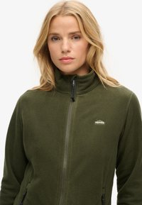 Olijfgroene fleece jas met een hoge kraag, ritssluiting en een logo aan de linkerkant. Voorzien van zijzakken en een zachte, gestructureerde stof.