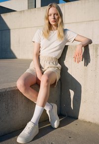 Junge Frau mit langen blonden Haaren, die ein weißes T-Shirt, beige Shorts, weiße Socken und Turnschuhe trägt, sitzt auf einer Betonkante im Freien im Sonnenlicht.