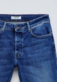 Tamne traperice tamnoplave boje s regularnim krojem, pet džepova, zakopčavanje na mesingasti gumb i žute šavove. Na oznaci piše "Salsa Jeans."