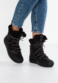 Bottes noires avec col en fausse fourrure, dessus en cuir texturé, semelle noire épaisse et lacets à motif. Conception hauteur cheville avec bout rond.