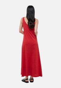 Robe longue rouge sans manches en tissu doux, avec une coupe décontractée, un design fluide et un décolleté arrondi dans le dos. Assortie à des sandales noires.