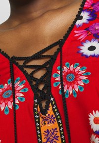 La tunica floreale rossa presenta un collo a V con lacci intrecciati neri, motivi floreali in rosa, blu e bianco, e dettagli intricati in nero.