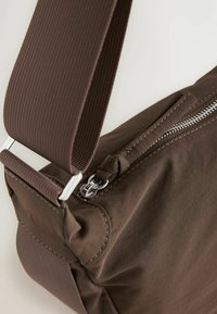 Brun skuldertaske i struktureret stof med en sølv lynlås, justerbar rem og stitched detaljer. Kompakt og funktionelt design.