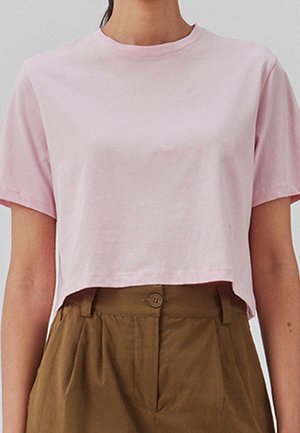 Korte, cropped t-shirt met korte mouwen van lichtroze katoen. Heeft een ronde halslijn en een relaxed fit, gecombineerd met hoge bruine broeken.