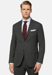 Blazer en gris sur mesure avec revers crantés, deux boutons et poches à rabat. Présente une chemise habillée bleu clair et une cravate bordeaux avec un mouchoir de poche.