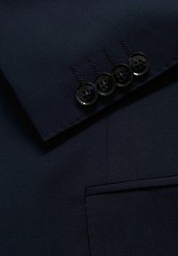 Marinblå kavaj med en texturerad yta, med slagkrage, fyra svarta knappar på ärmen och en sidficka.