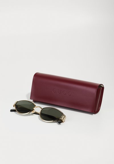 Slnečné okuliare Gucci s oversized oválnymi šošovkami, rámom so zlatými akcentami a čiernymi detailami na stranách. Súčasťou je bordová kožená etui s embosovaným nápisom "Gucci."