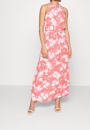 Vrouw draagt een koraalkleurige maxi-jurk met halternek en witte bloemenprint, met een gestrikte taille, gecombineerd met witte sandalen met hoge hakken, staand tegen een witte achtergrond.