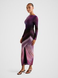 Moteris dėvi ilgomis rankovėmis purpurinę ir rožinę ombre su raukiniais suknią su priekine skeltuke, dėvi rudos spalvos aukštakulnius sandalus, stovinti ant pilkos grindų dangos.