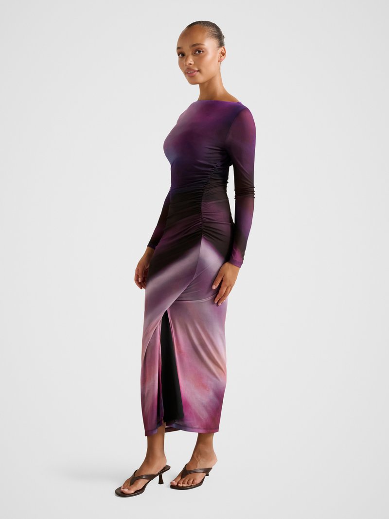 Moteris dėvi ilgomis rankovėmis purpurinę ir rožinę ombre su raukiniais suknią su priekine skeltuke, dėvi rudos spalvos aukštakulnius sandalus, stovinti ant pilkos grindų dangos.