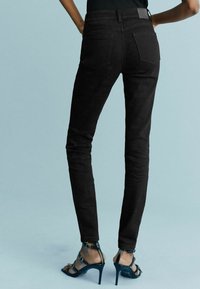 Personne portant un jean slim noir et des sandales à talons hauts noirs, debout devant un fond bleu clair.