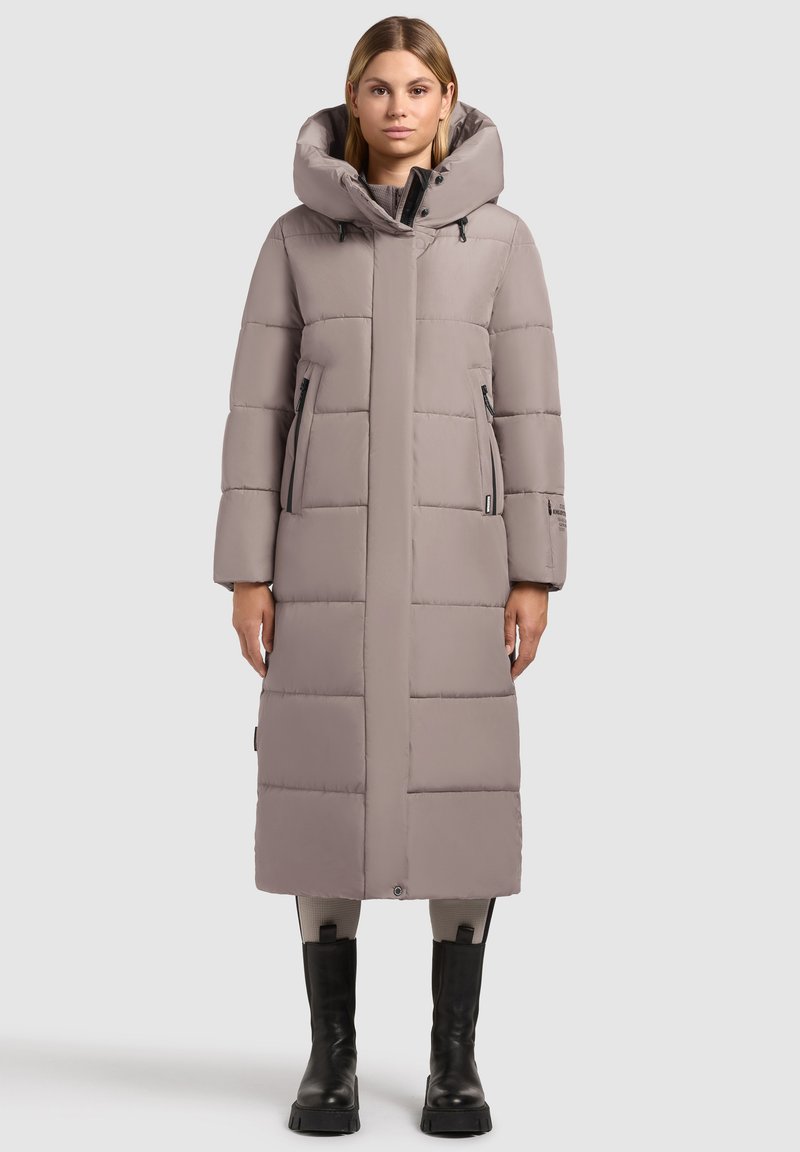 Lang, lys grå dunjakke med hætte, lynlåslommer i siderne og en sømdesign. Glat tekstur, knælang, parret med sorte støvler.