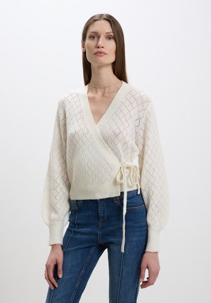 Cardigan en tricot crème avec motif en losanges, manches bouffantes longues et lien à la taille. Associé à un jean en denim bleu taille haute.