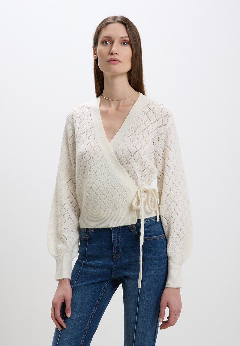 Maglioncino avvolgente lavorato a maglia color crema con motivo a diamante, maniche lunghe e voluminose e cintura in vita. Abbinato a jeans denim blu a vita alta.