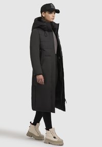 Duga crna puffer jakna s kapuljačom, koja sadrži kreširan dizajn i dva bočna džepa. Nosena preko uskih crnih tajica i bež čizama sa debelim đonom.