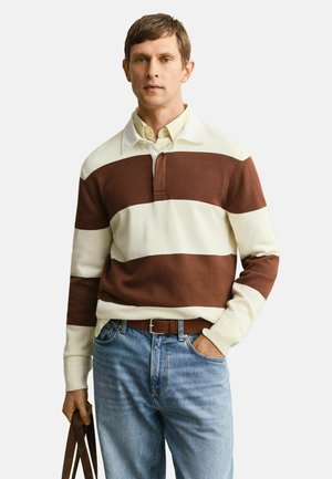 Mann trägt ein braun-weiß gestreiftes Langarmshirt über einem gelben Hemd mit Kragen, blaue Jeans und hält eine braune Umhängetasche.