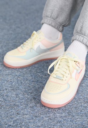 Osoba nosząca pastelowo żółte, różowe i niebieskie sneakersy Nike Air, szare spodnie dresowe i białe skarpetki na niebieskim wykładzinowym podłodze.