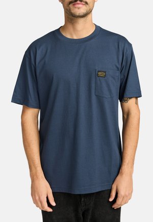 Man wearing navy blue short-sleeve t-shirt with chest pocket featuring a small black and yellow RVCA logo patch.