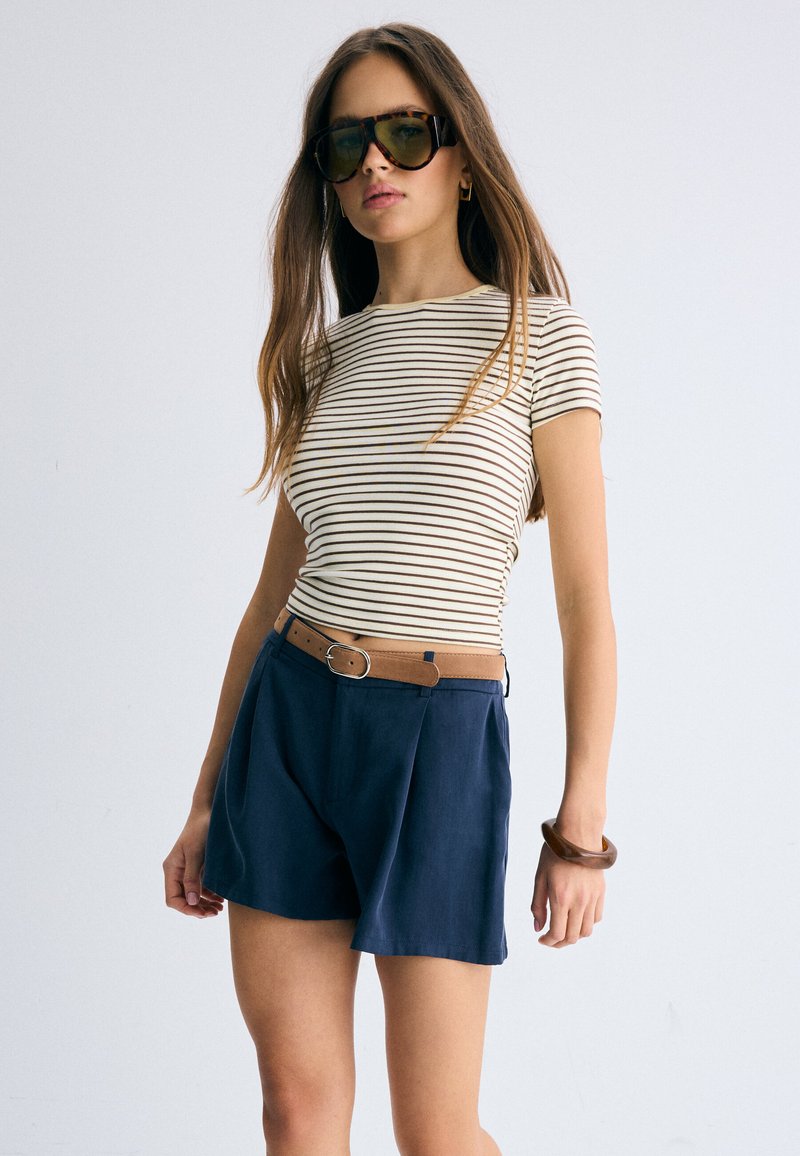 Femme portant de grandes lunettes de soleil, un haut à manches courtes rayé blanc et marron, un short taille haute bleu marine avec une ceinture marron, et un bracelet marron.