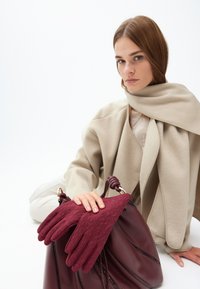 Burgundijske teksturirane rukavice odmaraju se na dubokoj burgundijskoj torbi s uvrnutim trakama, u kombinaciji s laganim bež šalom i svijetlom bluzom.