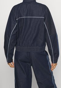 Person som bär en mörkblå denimjacka och matchande byxor med ljusblå sidostråkar, visad från baksidan mot en enfärgad bakgrund.