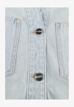 Lichtblauwe denim stof met twee metalen knopen gegraveerd met "NUKUS" en zichtbare stiksels rondom twee rechthoekige zakken.