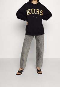 Felpa nera con cappuccio e lettering dorato "KORS", abbinata a jeans grigi a vita alta e sandali neri con tacco. Stile casual e sportivo.