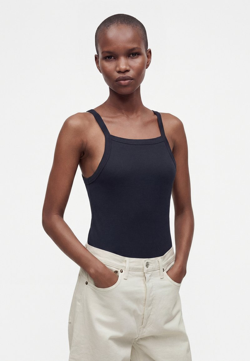 Femme aux cheveux courts portant un top noir moulant sans manches et un pantalon taille haute beige clair, les mains dans les poches, debout devant un fond uni.