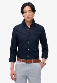 Camisa de botones azul marino con cuello, mangas largas y un bolsillo en el pecho. Combinada con pantalones de color claro y un cinturón de cuero marrón.