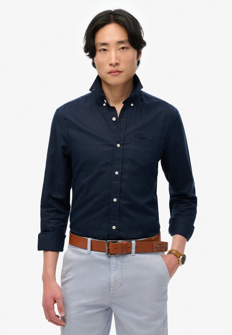 Camisa de botones azul marino con cuello, mangas largas y un bolsillo en el pecho. Combinada con pantalones de color claro y un cinturón de cuero marrón.
