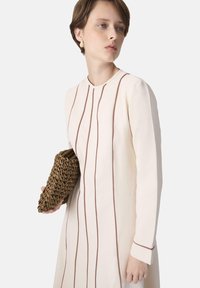 A young woman with short hair wears a cream dress with brown stripes, holding a woven brown clutch in her left hand.