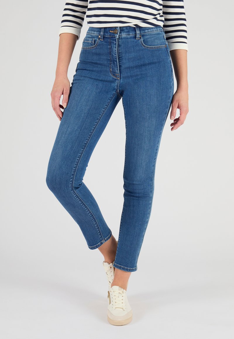 Jean en denim bleu taille haute avec une coupe slim, présentant des poches avant et une fermeture éclair. Modèle portant des baskets blanches.