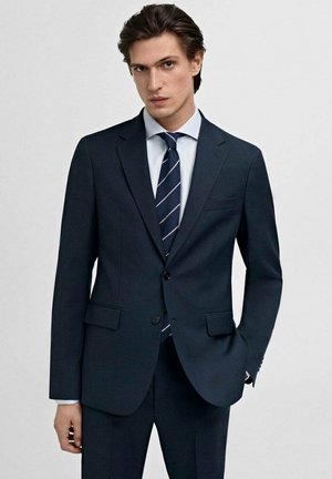 Costum bleumarin închis cu un design slim-fit, având două buzunare frontale și rever cu colțuri. Asortat cu o cămașă albă cu nuanțe de albastru deschis și o cravată în dungi.
