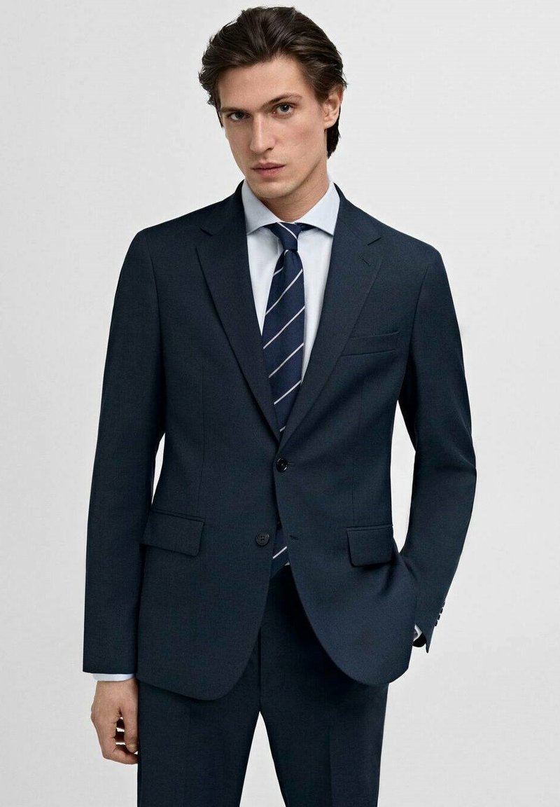 Ciemnoniebieski garnitur o slim-fit designie, z dwoma przednimi kieszeniami i klapami typu notch. Zestawiony z jasnoniebieską koszulą i wąskim krawatem w paski.